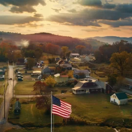 an American flag flies over a small town in the country's heartland with rolling hills and a setting sun in autumn