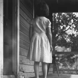 a young girl seen from behind stands on the porch of a humble house during the Great Depression