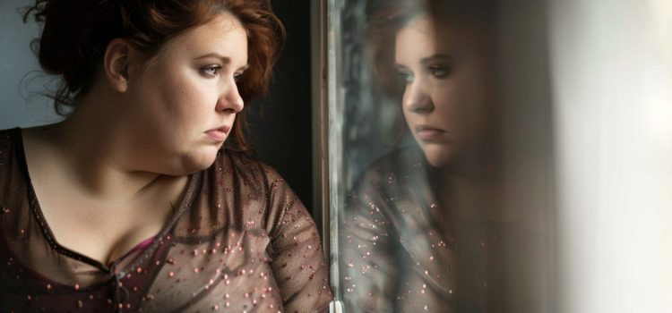 a sad overweight woman looking in a mirror raises the question, Why is body shaming bad?