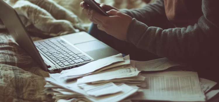 A person texting with their laptop open and papers next to them, showing one of the reasons why we procrastinate