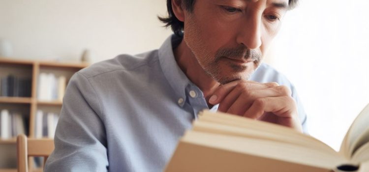 A middled-aged bearded man reading a book with shelves of books behind him