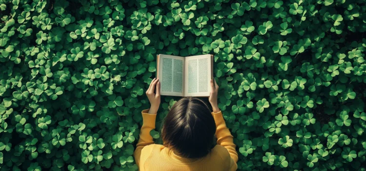 A girl wearing a yellow shirt reading a book while lying in a field of lucky four-leaf clovers