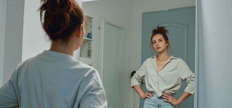 A woman looking in the mirror and her hands on her hips depicts the question "What is self-esteem, and why is it important?"