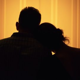 An image of a couple seen in silhouette from behind with the woman resting her head on the man's shoulder