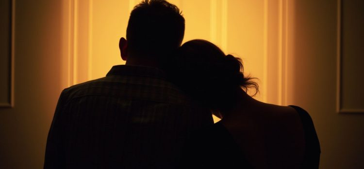 An image of a couple seen in silhouette from behind with the woman resting her head on the man's shoulder