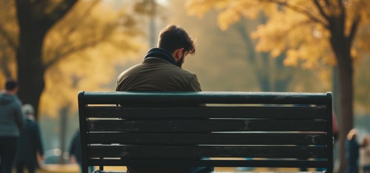 A sad man sitting on a park bench while other people walk by illustrates the question, "What is complicated grief?"