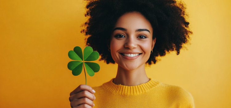 A smiling woman in a bright yellow sweater holding a large four-leaf clover illustrates the role of luck in success